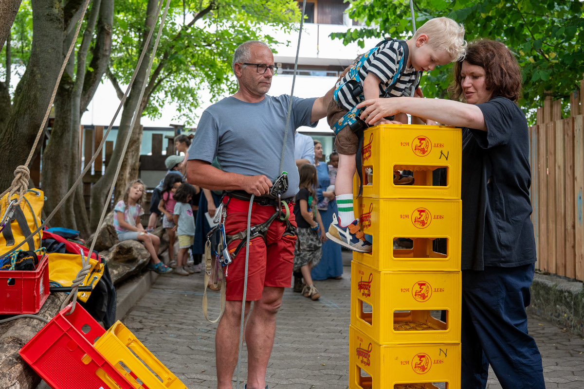 Ein kleiner Junge klettert mit Seilen gesichert an aufeinandergestapelten Getränkekästen empor. Eine Frau und eine Mann helfen ihm dabei.