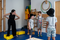 Eine Theaterpädagogin macht Kindern vor, wie sie einen griesgrämigen Riesen spielen können. Ein Kind spielt mit, das andere lacht die Pädagogin an. Ein drittes Kind steht mit dem Rücken zur Kamera gedreht da.