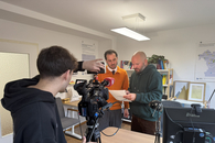Zwei Mitarbeiter stellen eine Szene nach, wie sie sich besprechen. Vor ihnen steht ein Mann mit Fernsehkamera. Er steht mit dem Rücken gedreht zum Fotografen.