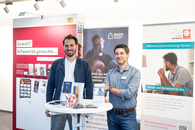 Zwei Projektleiter stehen vor Roll-ups ihrer Einrichtungen an einem Stehtisch mit Infomaterial.