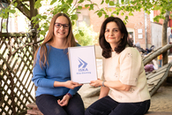 Die beiden Übergangsbegleiterinnen, eine dunkelhaarige Frau und eine etwas jüngere Frau mit Brille und blonden Haaren und Brille, sitzen unter einem Baum und halten ein Schild mit dem Projekttitel und dem ISKA-Logo in der Hand und lächeln in die Kamera.