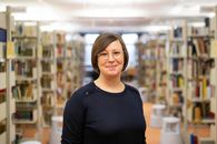 Die Referentin des beschriebenen Vortrags, eine Frau mittleren Alters mit Brille und kinnlangen braunen Haaren, steht in einer Bibliothek und lächelt in die Kamera.
