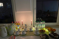 Ein angerichteter Tisch am Fenster mit Teekannen, Teetassen, Gebäck, Mandarinen und kalten Getränken. Draußen ist es bereits dunkel, zwei Kerzen brennen auf dem Tisch.