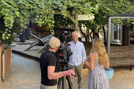 Eine Mann steht in einem Innenhof. Er wird von einer Frau im Sommerkleid interviewt, ein Kameramann steht daneben.