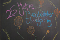 Tafel, auf der 25 Jahre Begleiteter Umgang steht. Drum herum sind Luftballons gemalt.