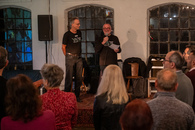 Zwei Männer stehen auf einer Bühne vor Publikum. Einer hält ein Mikrofon und ein Blatt Papier in der Hand.