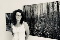 Schwarz-weiß-Foto einer Frau mit schwarzen Locken und weißem Top vor einem Foto an der Wand.