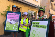 Ein Mann und zwei Frauen stehen im Freien vor einem Haus. Der Mann und eine der Frauen tragen Plakate am Körper, die zweite Frau hält Flyer in der Hand.