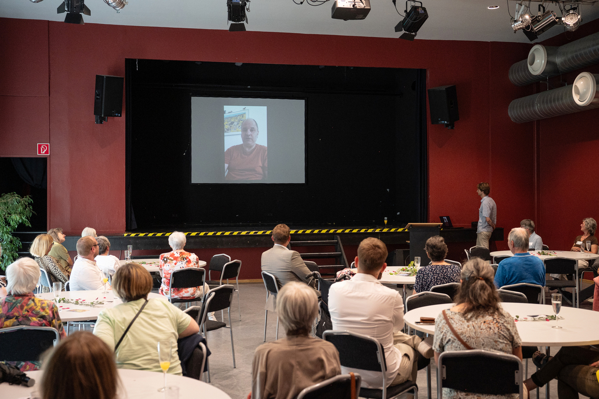 Videobotschaft von Egersdörfer auf einer Leinwand, davor Publikum.