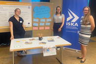 Drei ISAK-Fachdienste präsentieren das Projekt ISAK am Infostand. Im Hintergrund stehen ein ISKA-Roll-up, ein ISAK-Poster und ein Plakat mit den Ergebnissen des Workshops: Was brauchen Kinder, um gesund aufzuwachsen?.