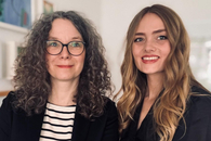 Zwei Frauen schauen freundlich in die Kamera, die linke ist etwas älter und hat braun-graues halblanges Haar, die rechte ist jünger und hat blondes langes Haar.
