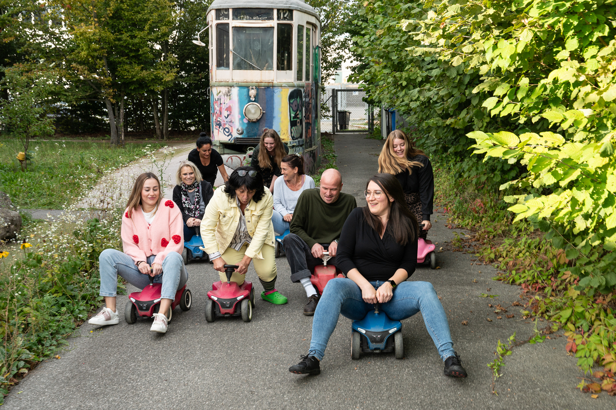 8 Frauen und ein Mann fahren auf Bobbycars und lachen in die Kamera