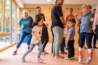 Kinder und Erwachsene stehen in oder rennen durch eine Turnhalle. Ein Kind springt vor Freude in die Luft.