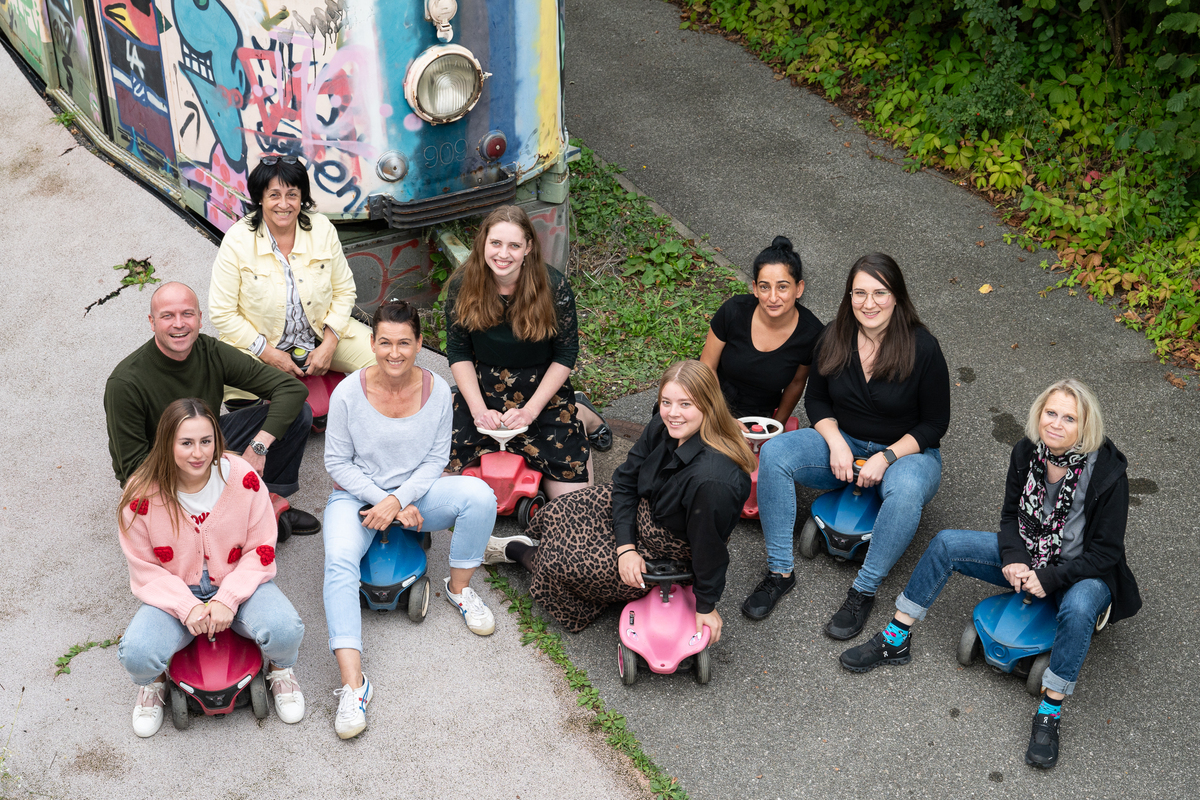 8 Frauen und ein Mann sitzen auf Bobbycars und lachen in die Kamera