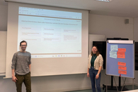 Ein Mann und eine Frau stehen lächelnd vor einer Wand, auf der eine Powerpoint-Präsentation projeziert ist. Am Rand steht ein Flipchart.