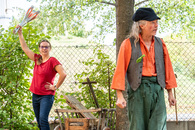 Ein bunt angezogener Mann steht im Garten. Im Hintergrund hält eine Frau Kegel in die Luft.