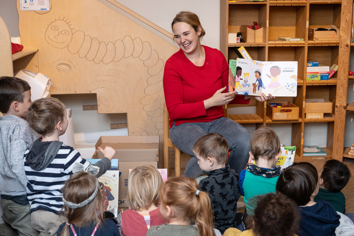 Eine Frau sitzt vor einer Kindegruppe und hält ein aufgeschlagenes Bilderbuch hoch.