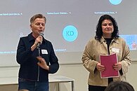 Eine Frau mit kurzen Haaren steht vor einem Publikum und spricht in ein Mikro. Sie hält einen Vortrag. Hinter ihr auf einer großen Leinwand sieht man Kacheln eines Videocalls, wodurch klar wird, dass es sich um eine hybride Veranstaltung handelt. Neben ihr steht eine dunkelhaarige Frau mit einem Papier in der Hand.
