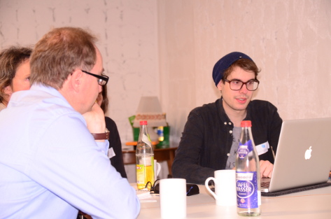 Ein Man mit einer Brille und einem blauen Hemd sitzt mit dem Rücken zur Kamera an einem Tisch. Er hört einem jüngeren Mann mit schwarzer Mütze und schwarzem Pulli zu, der ihm etwas erklärt.