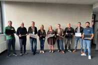 Neun Personen stehen vor einer Wand, jede Person hat einen aufgeklappten Laptop in der Hand.