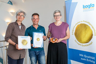 Zwei Frauen und ein Mann stehen zusammen für ein Gruppenfoto da. Die Frau links, bagfa-Vorsitzende, hat eine Plakette in der Hand, das QM-Siegel, das man an eine Wand anbringen kann. Der Mann, die Leitung des ZAB, hat die Urkunde in den Händen, die Frau rechts hält ein kleines Lebkuchenherz in den Händen. Rechts im Bild ist ein Banner der bagfa zum Thema QM-Siegel.
