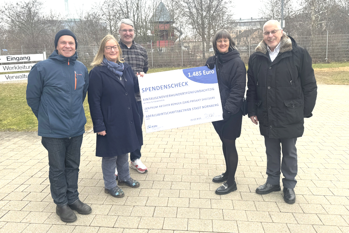 5 Personen stehen im Freien auf einer gepflasterten Fläche. 2 Personen in der Mitte halten einen großen symbolischen Spendenscheck in den Händen.