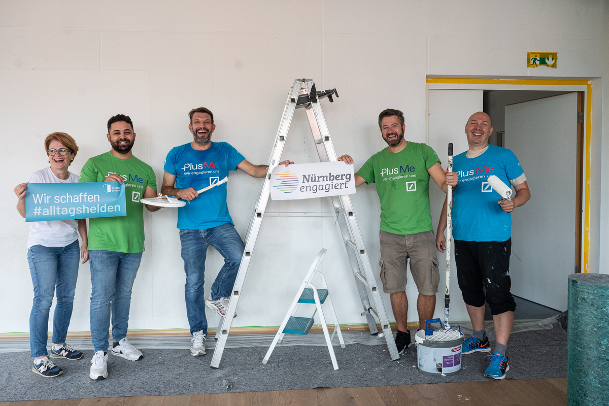 5 Personen mit Malerutensilien stehen neben einer Klappleiter, eine Person hält ein Schild mit der Aufschrift "Wir schaffen Alltagshelden",auf dem Boden ist Malervlies ausgelegt, darauf steht ein Farbeimer.